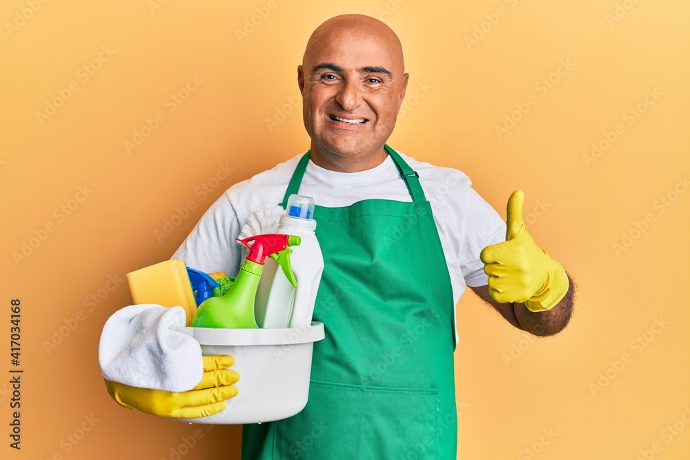 Mature middle east man wearing cleaner apron holding cleaning products smiling happy and positive, thumb up doing excellent and approval sign Mature middle east man wearing cleaner apron holding cleaning products smiling happy and positive, thumb up doing excellent and approval sign