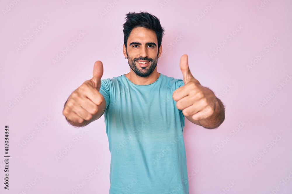 Handsome hispanic man with beard wearing casual clothes approving doing positive gesture with hand, thumbs up smiling and happy for success. winner gesture. Handsome hispanic man with beard wearing casual clothes approving doing positive gesture with hand, thumbs up smiling and happy for success. winner gesture.