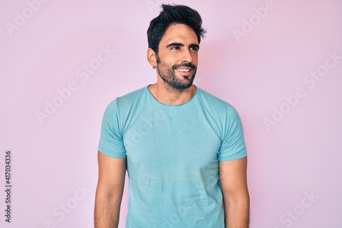 Handsome hispanic man with beard wearing casual clothes looking away to side with smile on face, natural expression. laughing confident.
