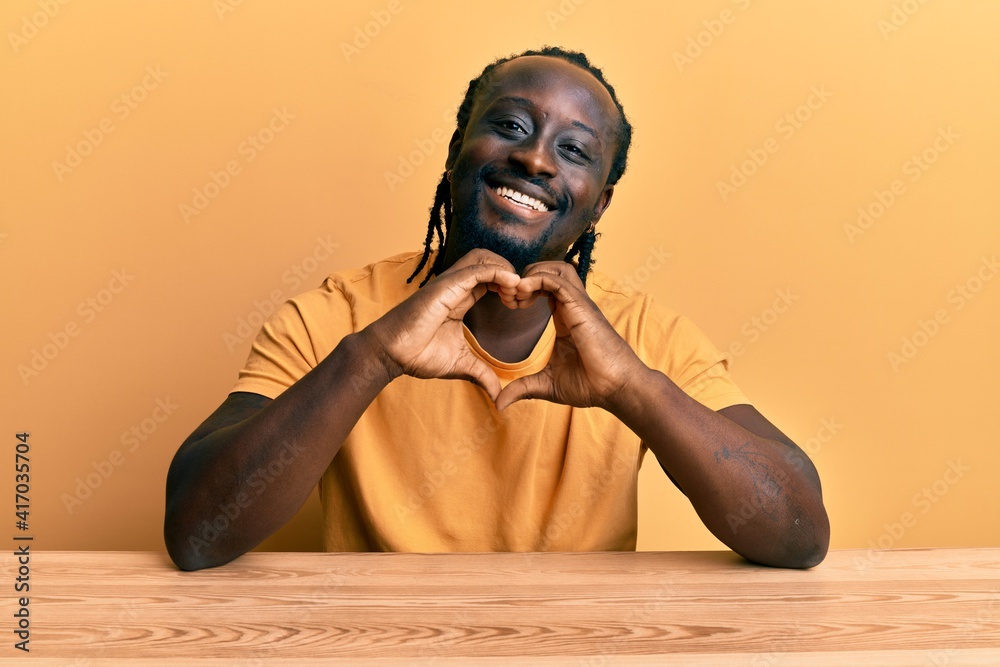 Handsome young black man wearing casual clothes sitting on the table smiling in love doing heart symbol shape with hands. romantic concept. Handsome young black man wearing casual clothes sitting on the table smiling in love doing heart symbol shape with hands. romantic concept.