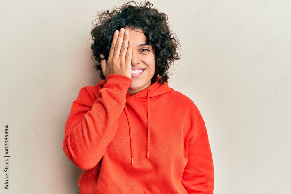 Young hispanic woman with curly hair wearing casual sweatshirt covering one eye with hand, confident smile on face and surprise emotion. Young hispanic woman with curly hair wearing casual sweatshirt covering one eye with hand, confident smile on face and surprise emotion.