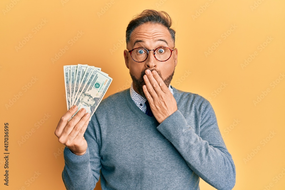 Handsome middle age man holding 20 dollars banknotes covering mouth with hand, shocked and afraid for mistake. surprised expression Handsome middle age man holding 20 dollars banknotes covering mouth with hand, shocked and afraid for mistake. surprised expression