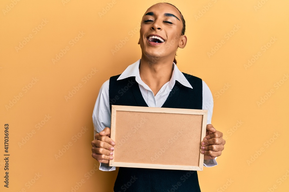 Hispanic man wearing make up and long hair holding empty corkboard smiling and laughing hard out loud because funny crazy joke. Hispanic man wearing make up and long hair holding empty corkboard smiling and laughing hard out loud because funny crazy joke.