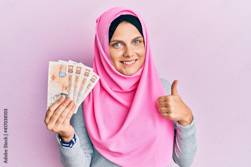 Young caucasian woman wearing islamic hijab holding 10 pounds banknotes smiling happy and positive, thumb up doing excellent and approval sign Young caucasian woman wearing islamic hijab holding 10 pounds banknotes smiling happy and positive, thumb up doing excellent and approval sign
