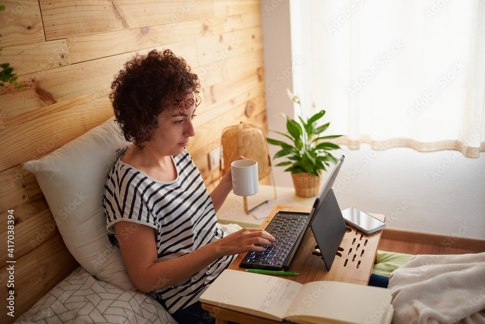 Woman telecommuting from her bed while holding a cup of coffee. Woman telecommuting from her bed while holding a cup of coffee.