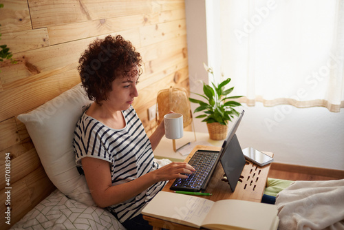 Woman telecommuting from her bed while holding a cup of coffee. photo