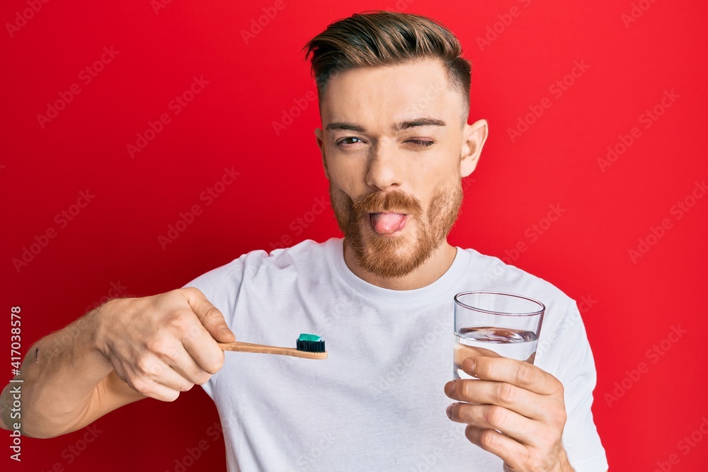 Young redhead man holding toothbrush with toothpaste and glass of water sticking tongue out happy with funny expression. Young redhead man holding toothbrush with toothpaste and glass of water sticking tongue out happy with funny expression.