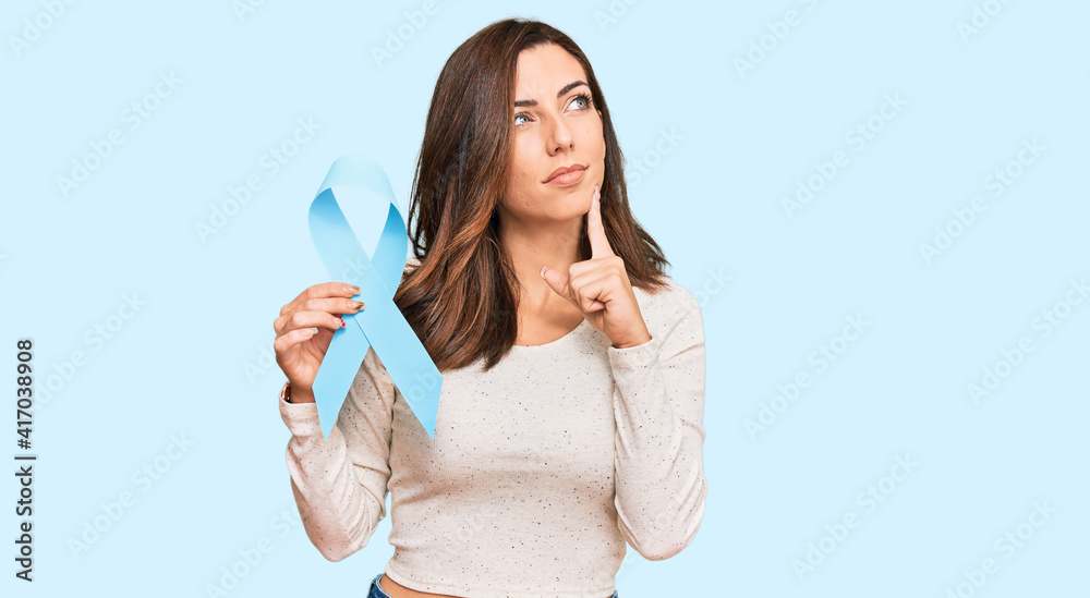 Young brunette woman holding blue ribbon serious face thinking about question with hand on chin, thoughtful about confusing idea Young brunette woman holding blue ribbon serious face thinking about question with hand on chin, thoughtful about confusing idea