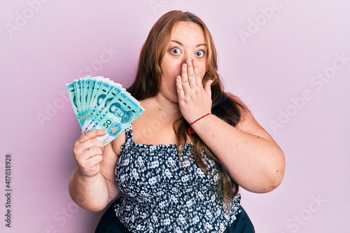 Plus size caucasian young woman holding 50 yuan chinese banknotes covering mouth with hand, shocked and afraid for mistake. surprised expression photo