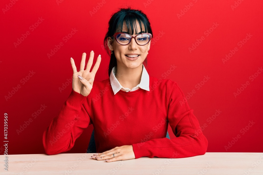 Young brunette woman with bangs wearing glasses sitting on the table showing and pointing up with fingers number four while smiling confident and happy. Young brunette woman with bangs wearing glasses sitting on the table showing and pointing up with fingers number four while smiling confident and happy.