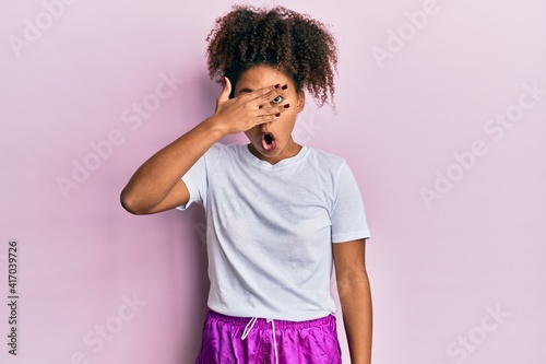 Beautiful african american woman with afro hair wearing sportswear peeking in shock covering face and eyes with hand, looking through fingers with embarrassed expression.