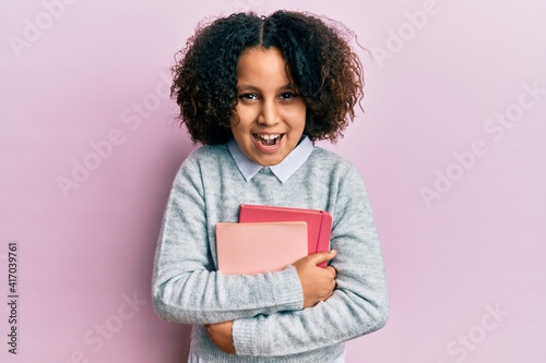 Young little girl with afro hair holding books smiling and laughing hard out loud because funny crazy joke.