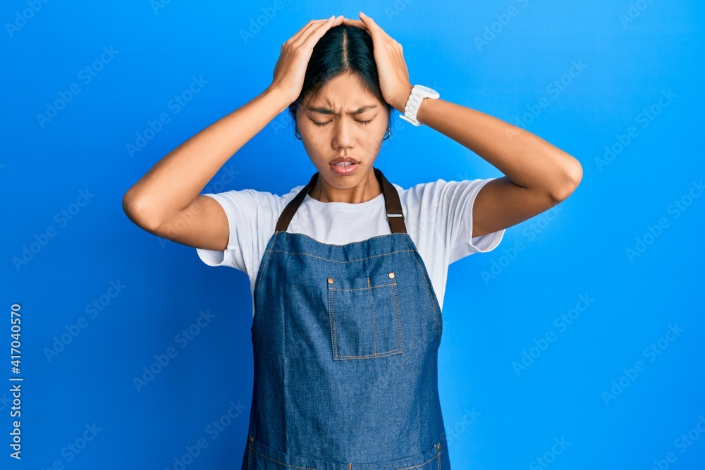Young chinese woman wearing waiter apron suffering from headache desperate and stressed because pain and migraine. hands on head.