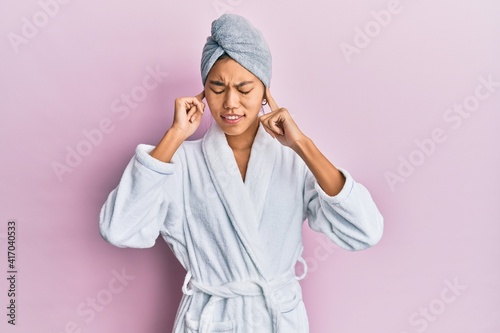 Young chinese woman wearing shower towel cap and bathrobe covering ears with fingers with annoyed expression for the noise of loud music. deaf concept. © Krakenimages.com