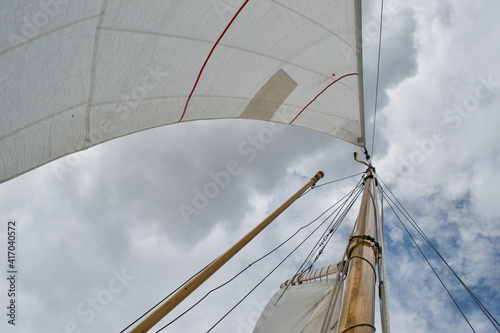 Looking up the mast of a gaff rigged sailing yacht: white sails, wooden mast and gaff, rope shrouds, halyards and sheets, and blue cloudy sunny sky