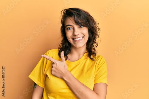Young hispanic woman wearing casual yellow t shirt smiling cheerful pointing with hand and finger up to the side