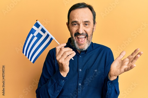 Middle age hispanic man holding greece flag celebrating achievement with happy smile and winner expression with raised hand