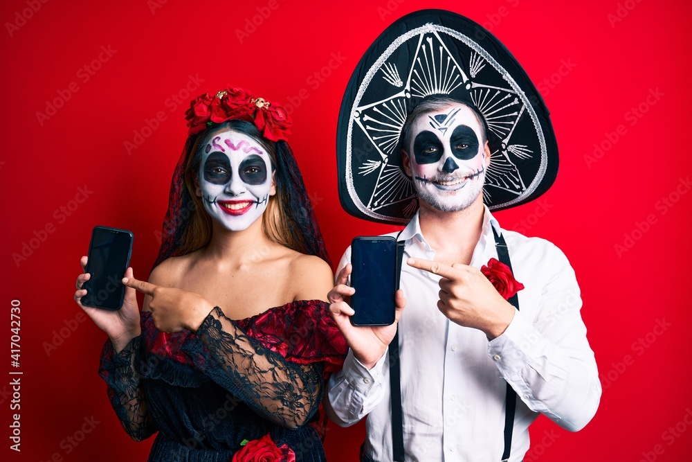 Couple wearing day of the dead costume holding smartphone showing screen smiling happy pointing with hand and finger Couple wearing day of the dead costume holding smartphone showing screen smiling happy pointing with hand and finger