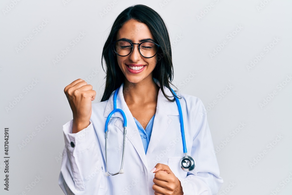 Beautiful hispanic woman wearing doctor uniform and stethoscope celebrating surprised and amazed for success with arms raised and eyes closed Beautiful hispanic woman wearing doctor uniform and stethoscope celebrating surprised and amazed for success with arms raised and eyes closed