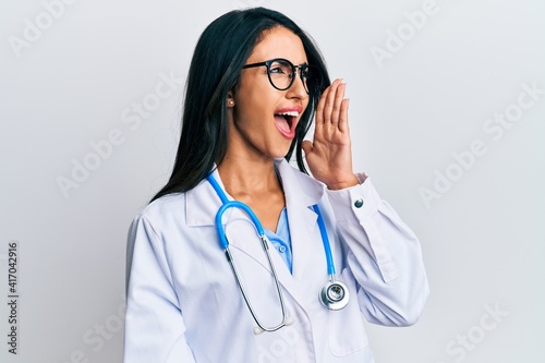 Beautiful hispanic woman wearing doctor uniform and stethoscope shouting and screaming loud to side with hand on mouth. communication concept.