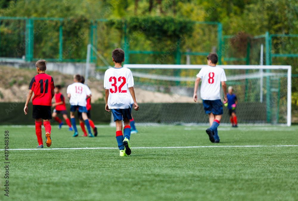 Obraz premium Young sport boys in white and red sportswear running and kicking a ball on pitch. Soccer youth team plays football in summer. Activities for kids, training 