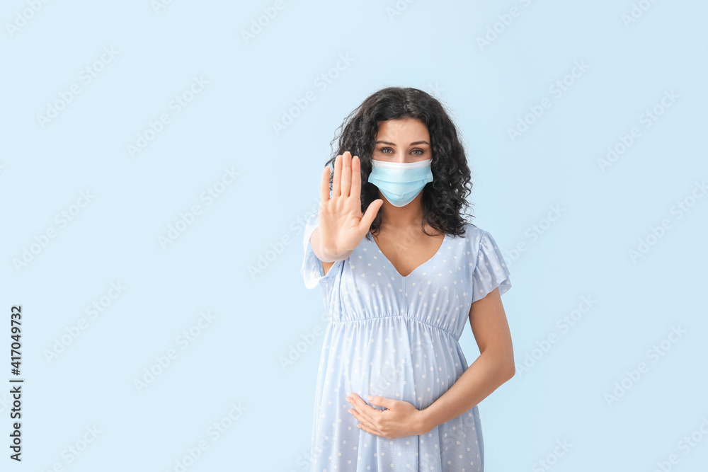Pregnant woman in medical mask showing stop gesture on color background