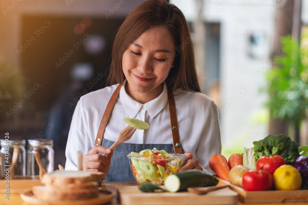 Closeup image of a beautiful young asian female chef cooking and eating fresh mixed vegetables salad in kitchen