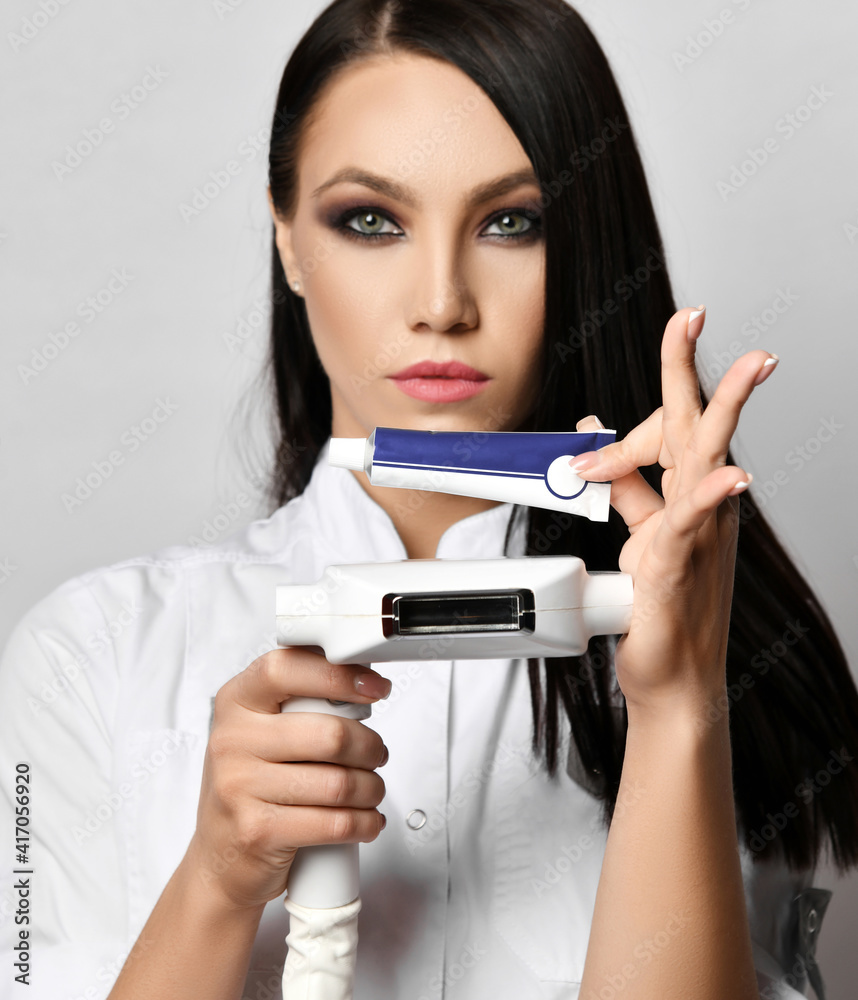 Woman doctor cosmetologist in medical gown holds equipment for laser hair  removal and tube with special remedy cream gel over grey wall background.  Beauty, skincare concept Stock Photo | Adobe Stock