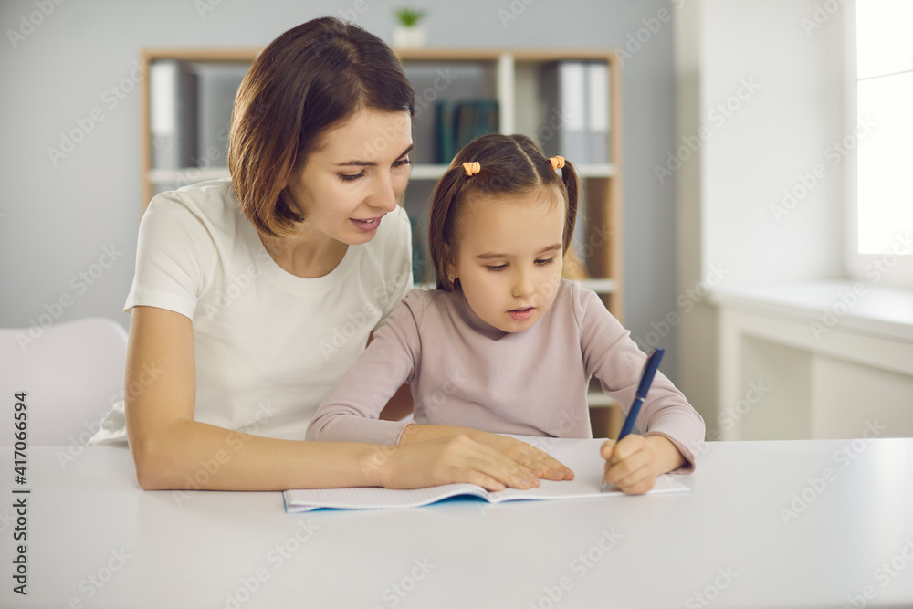 Small concentraited attentive girl sitting with mother or nanny together in room and drawing or learning alphabet and writing. Children development and education, activities with kids concept Small concentraited attentive girl sitting with mother or nanny together in room and drawing or learning alphabet and writing. Children development and education, activities with kids concept