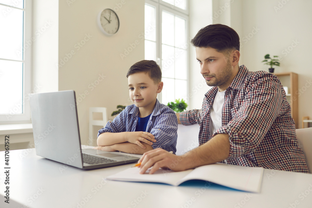 Father helping son in doing homework. Caring dad sits at a table with his son and helps him with his studies using a laptop to study school lessons. Concept of education, e-learning and parenting. Father helping son in doing homework. Caring dad sits at a table with his son and helps him with his studies using a laptop to study school lessons. Concept of education, e-learning and parenting.