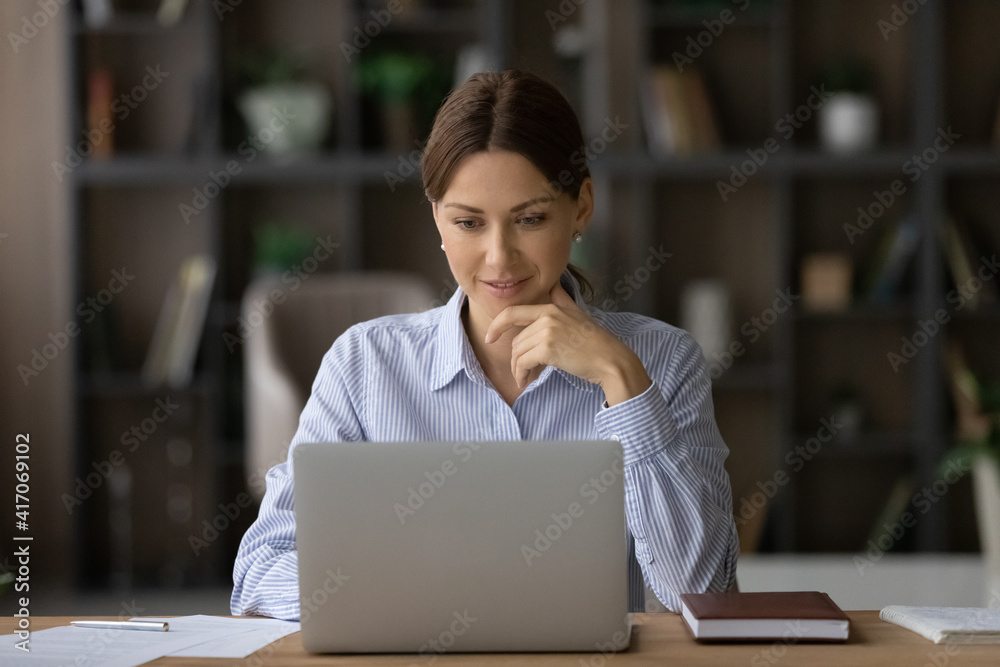 Fototapeta premium Smiling young Caucasian businesswoman sit at desk at home office look at laptop screen consult client online. Millennial female employee or woman worker work distant on computer, analyze paperwork.