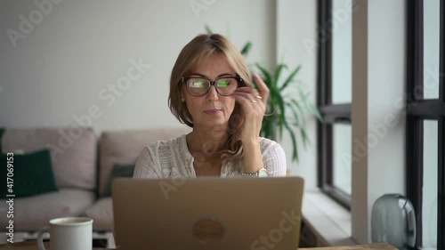 Businesswoman working with laptop and sitting at table in home office during pandemic. Front view of middle aged American woman looks at computer screen and types text, sits at desk in light room with