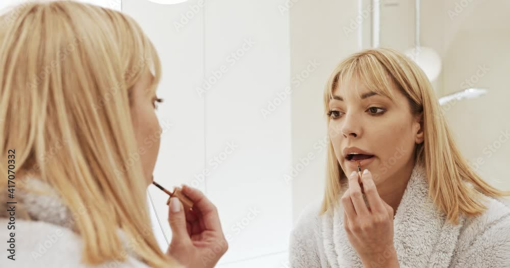 Morning makeup. The woman paints her lips with lipstick. A young woman dressed in a bathrobe paints her lips in front of the bathroom mirror. Morning makeup. The woman paints her lips with lipstick. A young woman dressed in a bathrobe paints her lips in front of the bathroom mirror.