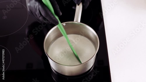 chef man in black gloves preparing a white liquid dessert in an iron pan on an induction stove stirring with a green spatula
