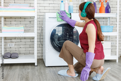 Housework woman listing music by headphone and cleaning washing machine in the laundry room.