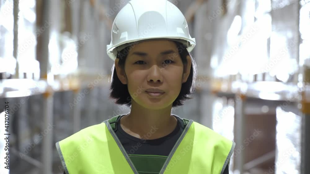 Portrait of asian woman with smile standing at warehouse, looking at camera. Happy warehouse worker at workplace. employee in rows of storage racks with merchandise. concept uniform Portrait of asian woman with smile standing at warehouse, looking at camera. Happy warehouse worker at workplace. employee in rows of storage racks with merchandise. concept uniform