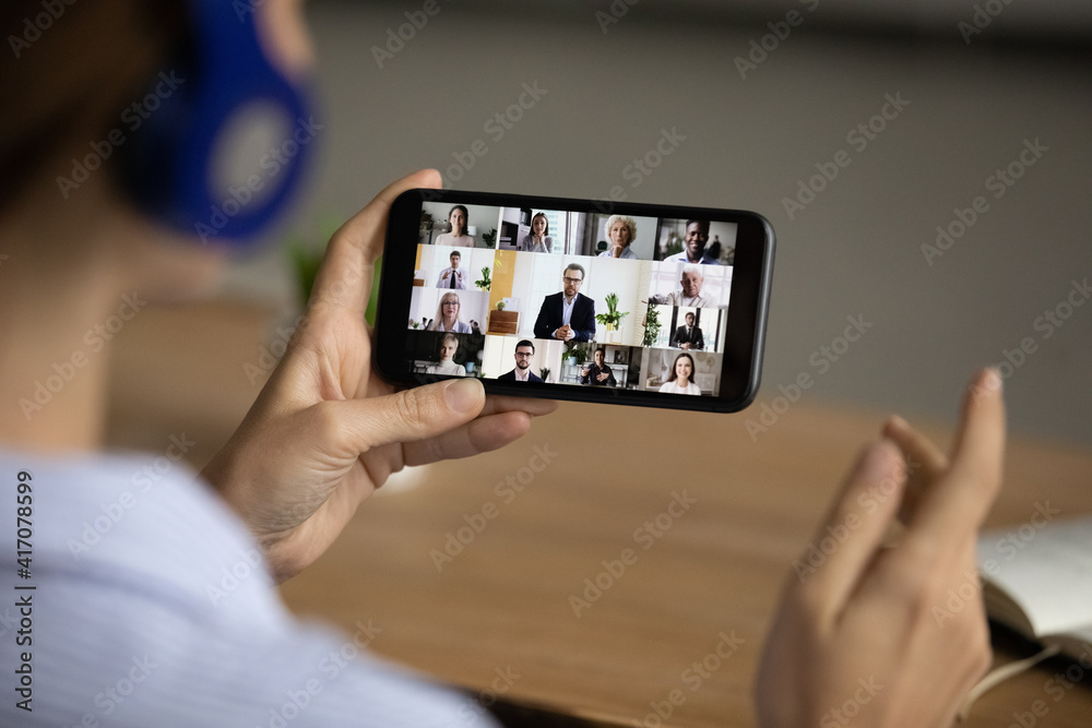 Over shoulder view of businesswoman have online webcam team meeting with colleagues on smartphone at home office. Woman talk speak on video call on cellphone with coworkers. Technology concept.