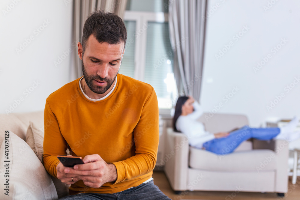 Husband turned his back to wife, reading message on phone from his lover, Woman lying on sofa in the back. Cheating and infidelity concept Husband turned his back to wife, reading message on phone from his lover, Woman lying on sofa in the back. Cheating and infidelity concept