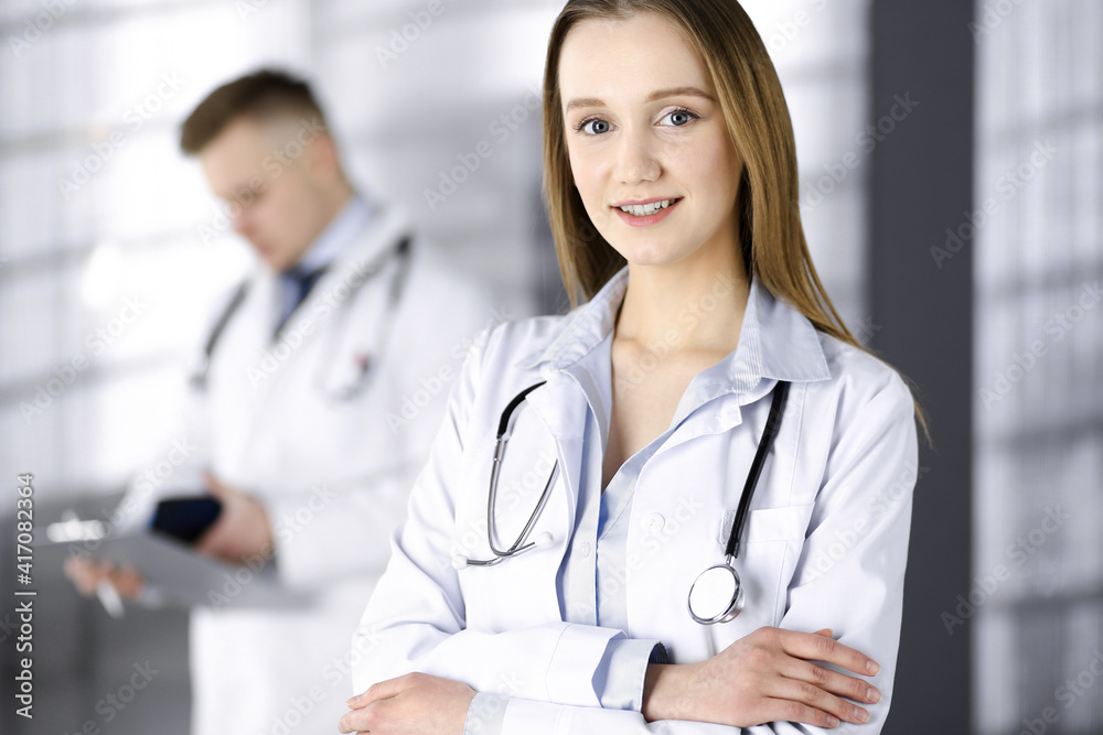 Professional beautiful woman-doctor with a stethoscope is standing with crossed arms in a clinic. Young doctors at work in a hospital. Medicine and healthcare concept Professional beautiful woman-doctor with a stethoscope is standing with crossed arms in a clinic. Young doctors at work in a hospital. Medicine and healthcare concept