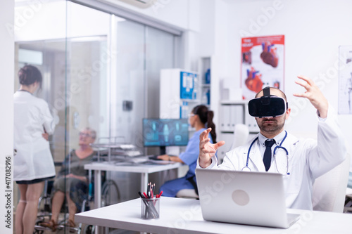 Portrait of professional caucasian male doctor in VR glasses sitting in hospital cabinet and making gestures using virtual reality innovations while female nurse working in background