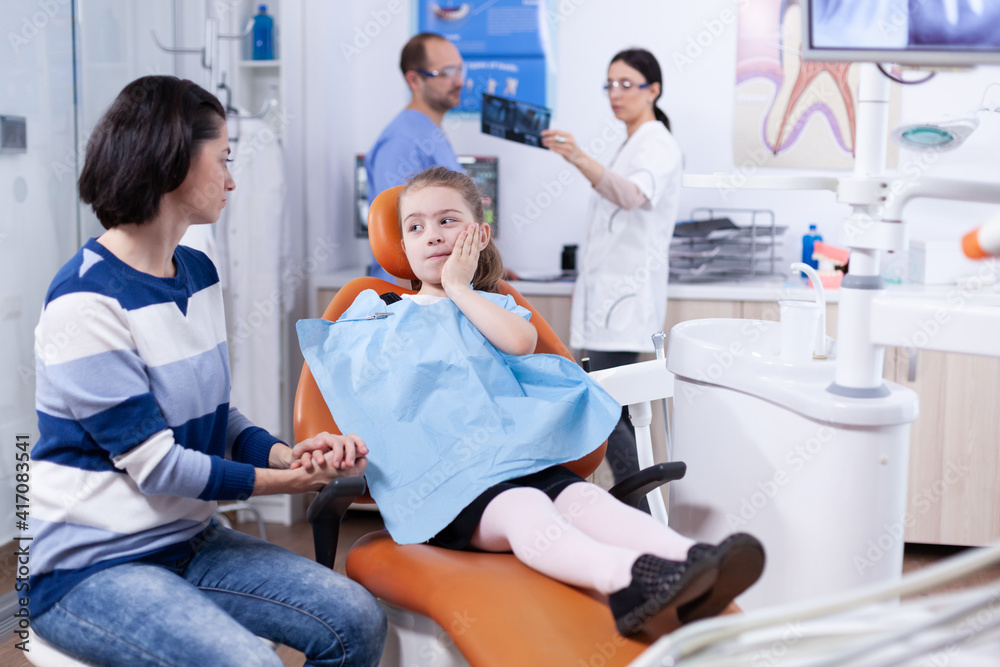 Little kid with painfull expression touching face during professional dental treatment. Child with her mother during teeth check up with stomatolog sitting on chair. Little kid with painfull expression touching face during professional dental treatment. Child with her mother during teeth check up with stomatolog sitting on chair.