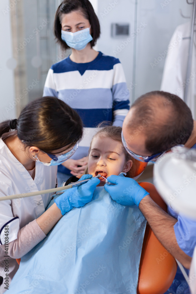 Dentist examining little girl's tooth with assistant help wearing uniform. Mother with her kid in stomatology clinic for teeth examine using modern instruments. Dentist examining little girl's tooth with assistant help wearing uniform. Mother with her kid in stomatology clinic for teeth examine using modern instruments.