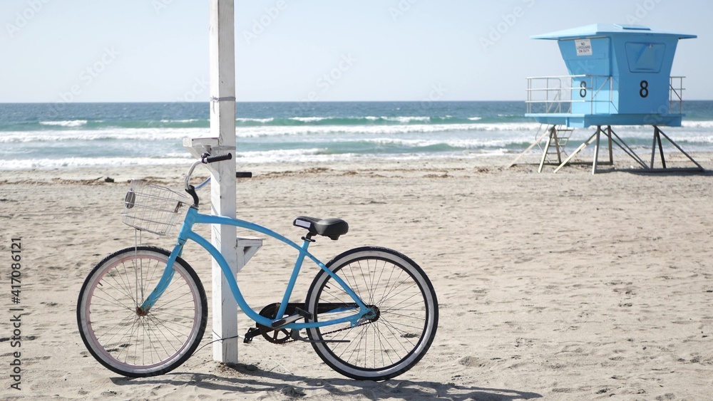 Fototapeta premium Blue bicycle, cruiser bike by ocean beach, pacific coast, Oceanside California USA. Summertime vacations, sea shore. Vintage cycle on sand near lifeguard tower or watchtower hut. Sky and water waves.