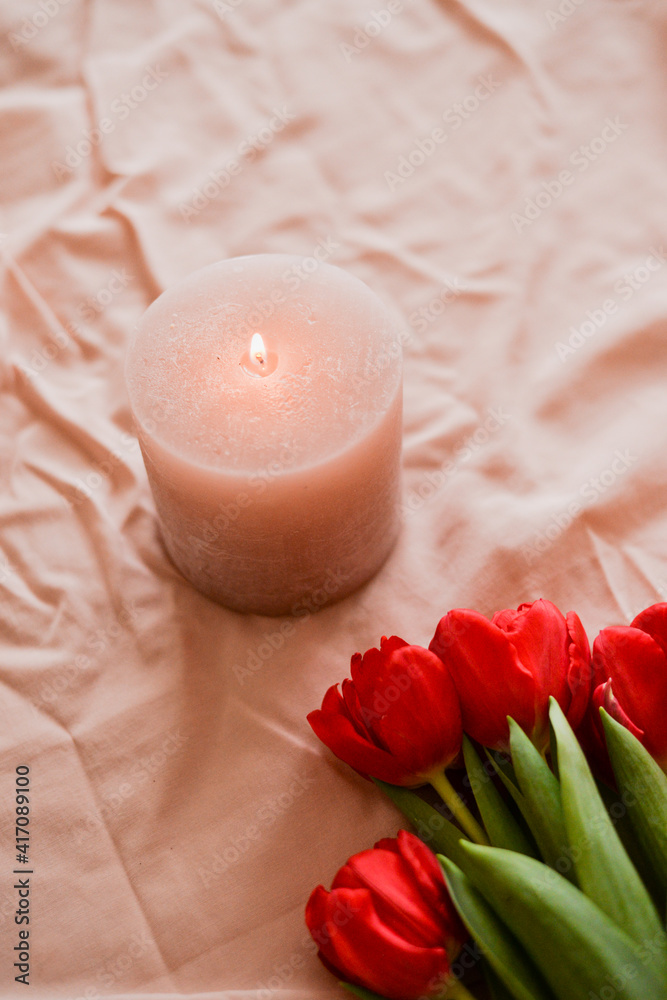 Pink candle with flame standing on the pink textile background and red tulips Pink candle with flame standing on the pink textile background and red tulips