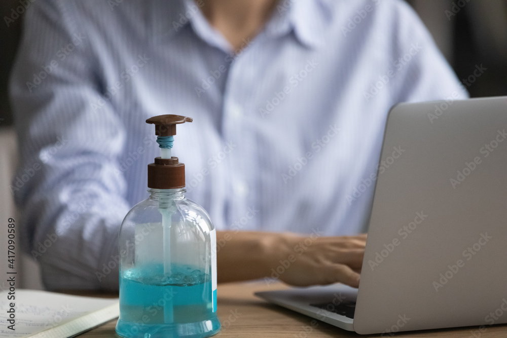 Close up of bottle with liquid sanitizer on desk in office, female employee work on computer in background. Antibacterial gel protect from coronavirus covid-19 pandemics at workplace. Corona concept.