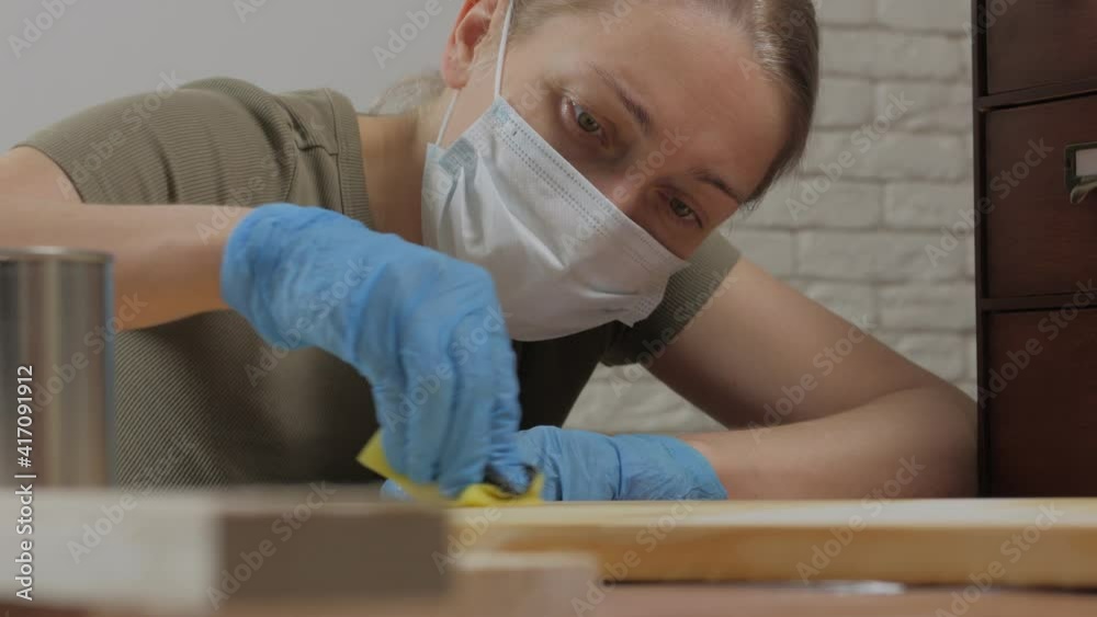 A female carpenter, designer, wearing a medical mask on her face, covers the wooden surface with oil. A female carpenter, designer, wearing a medical mask on her face, covers the wooden surface with oil.