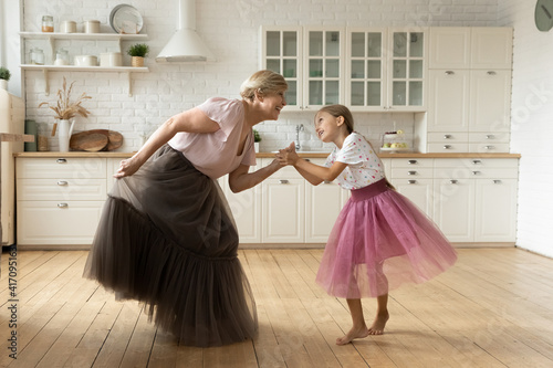 Active mature grandma dance with small granddaughter on warm floor at modern kitchen in puffy ballerina skirts. Joyful senior nanny play princesses with little girl dancing listen to music having fun