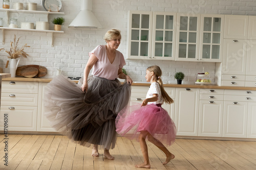 Merry leisure time. Happy energetic grandmother teach ball dance active little child. Caring grandma junior girl grandkid engaged in dancing in funny large fluffy skirts barefoot on warm kitchen floor photo