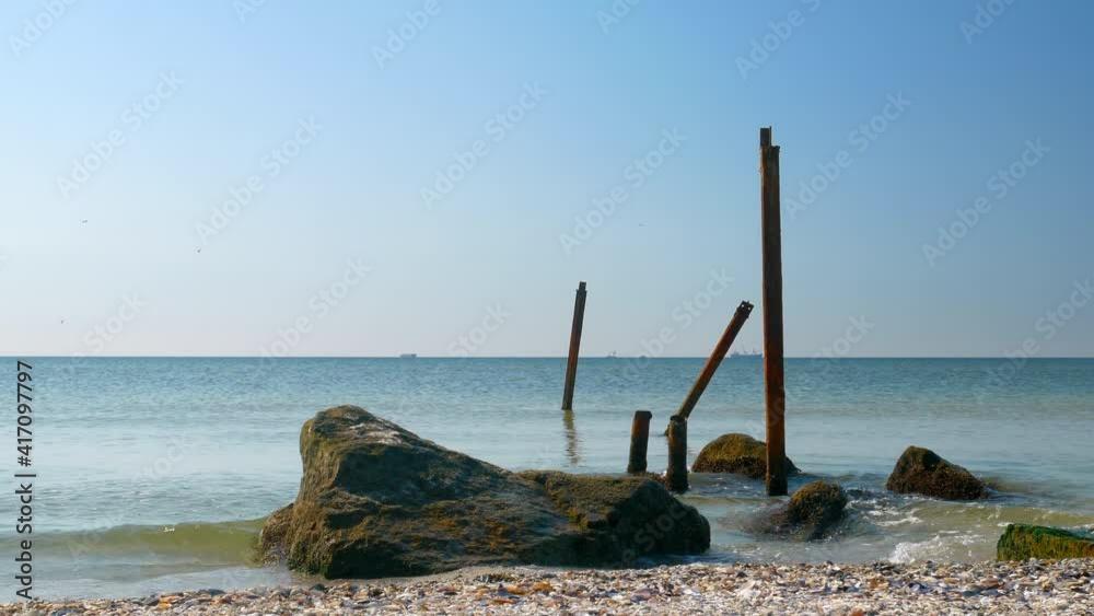 Close-up of the rusty wreckage of an old pier on the beach. Quiet gentle sea and clear blue sky around. Ships are at anchor on the horizon. Close-up of the rusty wreckage of an old pier on the beach. Quiet gentle sea and clear blue sky around. Ships are at anchor on the horizon.