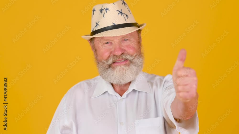 Happy elderly gray-haired bearded man in white shirt and hat showing thumb up. People lifestyle concept. Old man posing isolated on yellow background. Happy elderly gray-haired bearded man in white shirt and hat showing thumb up. People lifestyle concept. Old man posing isolated on yellow background.